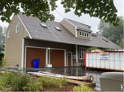 Large suburban house with multiple gables and brown garage doors, viewed in rain beside a construction dumpster and wet driveway.