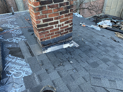 Urban rooftop with a brick chimney, gray shingles, graffiti-covered roofing membrane, scattered debris, and bare branches in the background.
