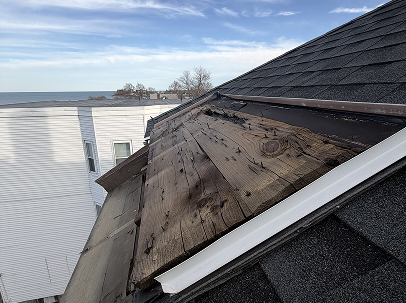 Severely damaged roof with exposed, weathered wooden decking and missing shingles; a quiet coastal neighborhood and blue sky appear behind it.