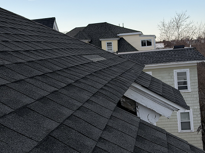 Close-up of overlapping dark gray asphalt roof shingles on a home, with white siding, windows, neighboring rooftops, and bare trees under a clear sky.