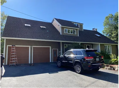 Sunlit suburban home with attached double garage, black SUV in driveway, dormer windows, green trees, and a clear blue sky.