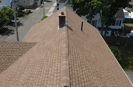 Aerial view of a suburban home’s brown shingle roof, brick chimney, skylight, neighboring houses, tree-lined yards, and quiet street.