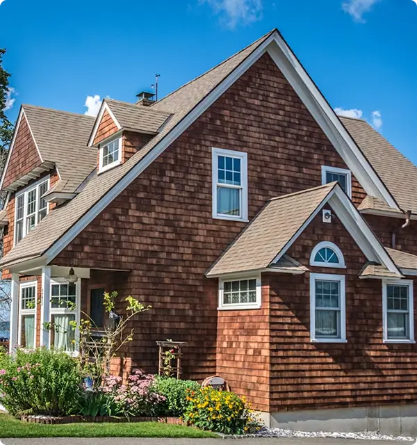 Sunlit Craftsman-style home with cedar-shingle siding, steep gables, white trim, blue sky, and colorful garden flowers creates a welcoming mood.
