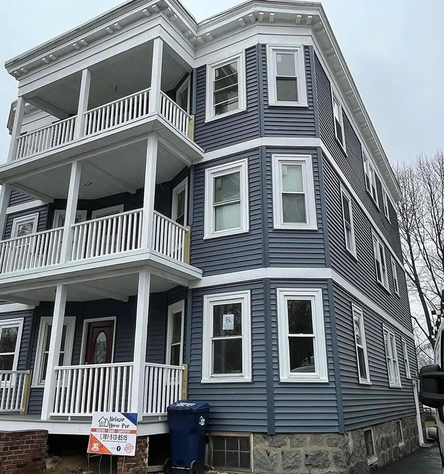 Three-story dark-blue Victorian home with white trim, stacked porches, bay windows, stone foundation, and a “For Sale” sign on a quiet street.