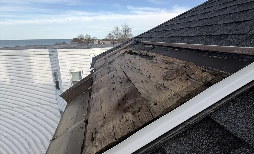 Close-up of a severely deteriorated roof with exposed, weathered wood and missing shingles, beside a white house overlooking calm water.