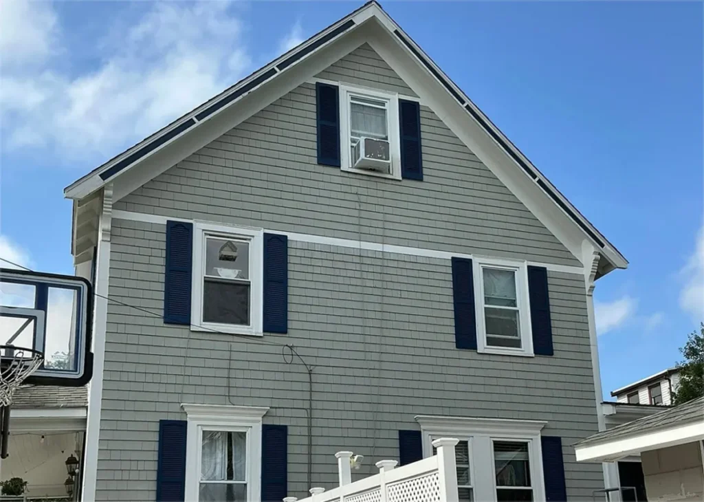 Gray-shingled house with navy shutters, white-trimmed windows, a basketball hoop, and porch under a bright blue, partly cloudy sky.